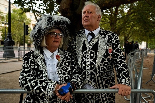 Un hombre y una mujer, vestidos con trajes brillantes, esperan el paso de la procesión fúnebre que se va a celebrar en Londres para despedir a Isabel II, que falleció hace más de una semana a los 96 años. AFP